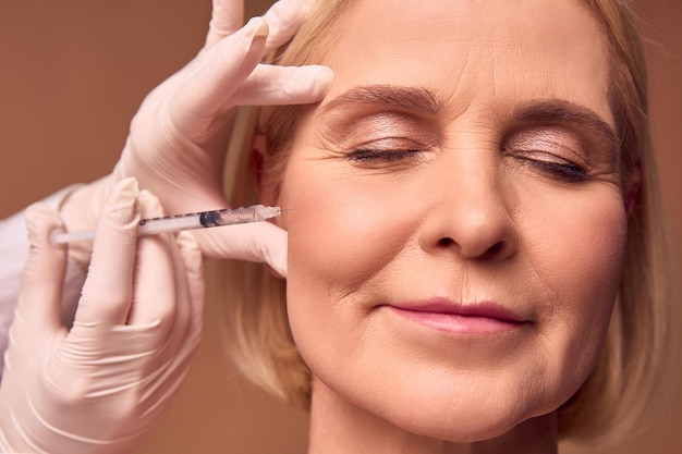 Foto close-up portret van een volwassen vrouw met gesloten ogen op een beige achtergrond handen in witte handschoenen en een medische jurk maken een injectie in het jukbeen het gladstrijken van rimpels esthetische geneeskunde