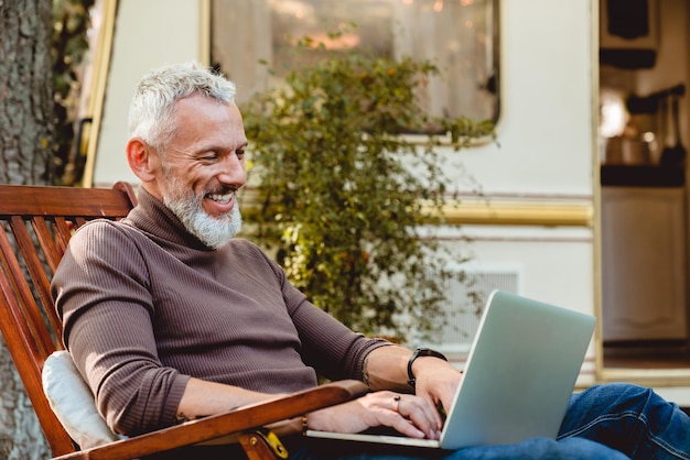 Close-up portret van een vrolijke oudere mannelijke reiziger die aan zijn laptop werkt in een leunstoel op de veranda van zijn caravan