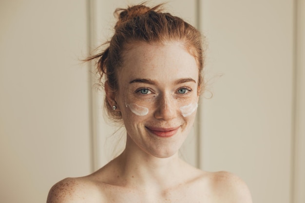 Close-up portret van een vrouw met rood haar en sproeten die crème op haar gezicht aanbrengt gezichtsbehandeling...