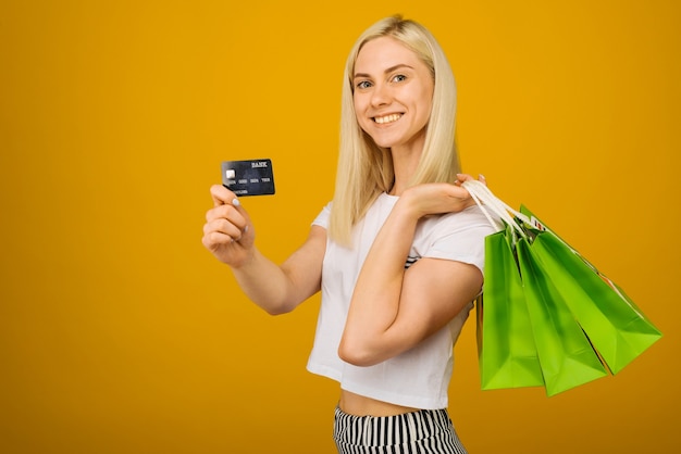 Close-up portret van gelukkige jonge mooie blonde vrouw met creditcard en groene boodschappentassen, camera kijken, op geel