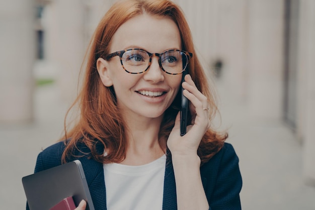 Close-up portret van glimlachende positieve roodharige zakenvrouw die een bril draagt die partner belt, mobiele telefoon in de ene hand houdt en laptop in de andere, iets bespreken op mobiele telefoon buitenshuis