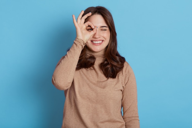 Close-up portret van jonge positieve vrouw met donker haar, mooie vrouw heeft speels goed humeur, die haar oog bedekt met ok teken, geïsoleerd over blauwe muur.