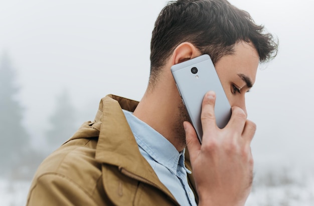 Foto close-up portret van knappe stijlvolle jonge blanke man met trendy jas naar beneden kijkend en pratend op zijn mobiele telefoon tijdens het wandelen op mistige lifestyle mensen en technologie concept