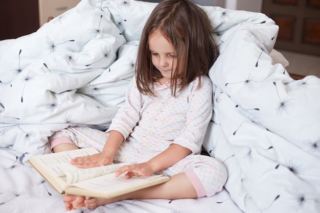 Close-up portret van meisje zittend in bed, het dragen van pyjama, charmante vrouwelijke jongen met donker haar, boek in de hand houden en het lezen, kind kijkt naar haar boek, leest interessant sprookje.