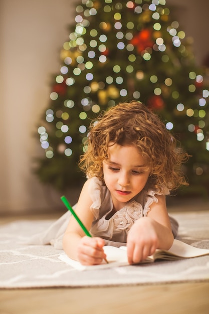 Close-up portret van mooi meisje met mooi krullend haar tijdens het schrijven van begroetingsmail aan haar grootouders op kerstavond in gezellige kamer in de buurt van dennenboom Gezellige wintervakantie concept