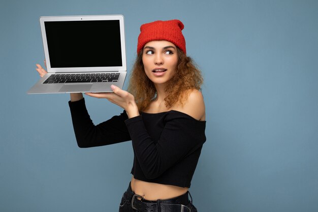 Foto close-up portret van mooie grappige charmante zelfverzekerde donkere blonde krullende jonge vrouw met laptopcomputer opzoeken met zwarte crop top en rode en oranje do-rag geïsoleerd over lichtblauwe w