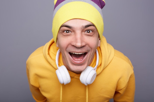 Close-up portret van opgewonden dolblije hipster man met gele hoodie en beanie hoed staande tegen grijze muur geluk uitdrukkend plotseling succes vierend naar camera kijkend wow