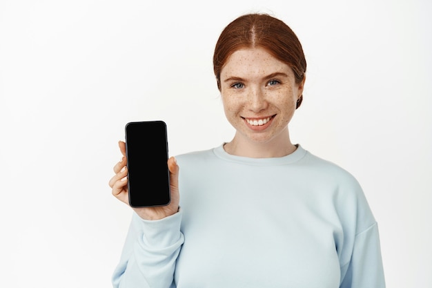 Close-up portret van roodharige meisje weergegeven: mobiele telefoon scherm en glimlachen. Jonge vrouw met mobiele telefoon op wit