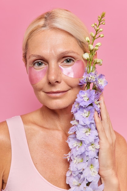 Close-up portret van serieuze vijftig jaar oude vrouw brengt hydrogel patches aan onder de ogen kijkt direct naar de camera houdt bloemen heeft een schone gezonde huid ondergaat schoonheidsprocedures poses binnen.