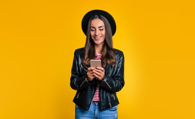 Close-up portret van stijlvolle moderne schattige hipster vrouw in zwarte jas en muts met slimme telefoon is surfen in het web terwijl ze geïsoleerd op gele achtergrond