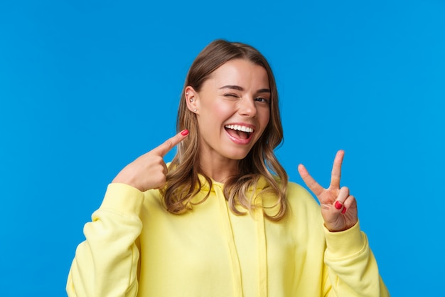 Close-up portret van trots en opschepperig schattig blond meisje met kort haar en doorboord oor, wijzend op zichzelf en vredesgebaar maken met een tevreden glimlach, staande blauwe muur vrolijk