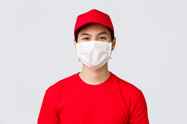 Close-up portret van vrolijke Aziatische bezorger in medische masker en rood GLB uniform, t-shirt, lachend met ogen naar de klant, het nemen van orde met veiligheidsmaatregelen om covid-19 verspreiding te voorkomen