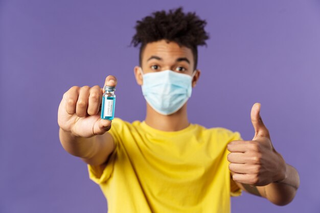 Close-up portret van vrolijke jonge Spaanse man in medische masker met ampul met coronavirus vaccin, coronavirus drug, show thumbs-up, beschoten ambulance, staande paarse achtergrond