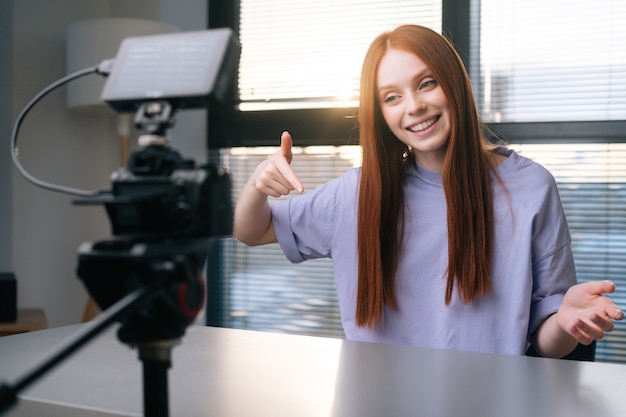 Close-up of positive young woman blogger talking about new phone using professional camera and softbox sitting at desk on background of window.