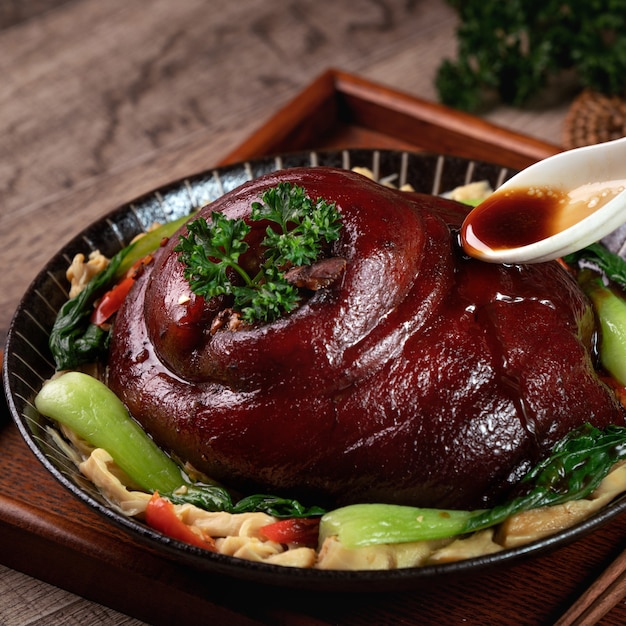 Закройте вверх наливания пикантного соевого соуса с ложкой над тайваньской едой тушеной свинины в тарелке на деревенском фоне стола.