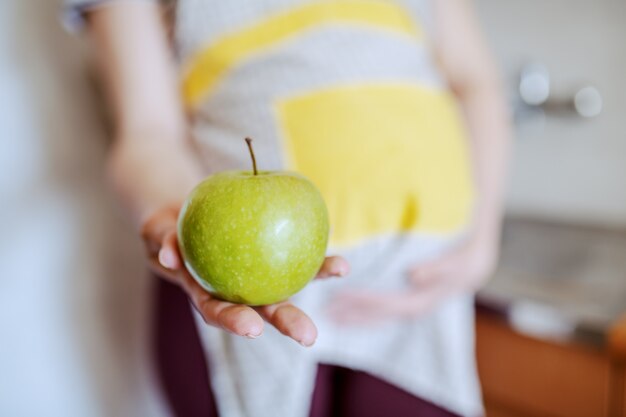 Close up of pregnant Caucasian woman in apron holding green apple. Selective focus on apple.