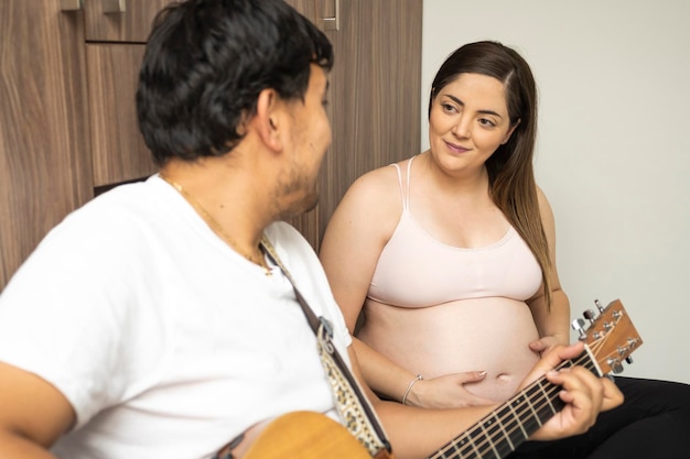 Close up pregnant wife looks at her husband with love while he is playing the guitar at home