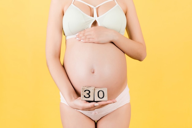 Крупным планом беременная женщина в красочном нижнем белье, держащая кубики с числами недель беременности. Тридцать недель беременности на желтом фоне. Скопируйте пространство.