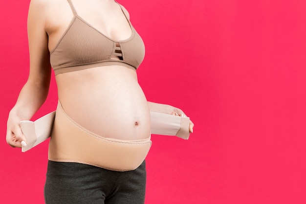 Close up of pregnant woman dressing elastic maternity band at pink surface with copy space. Orthopedic abdominal support belt concept.