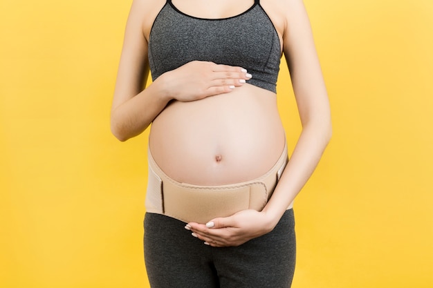 Close up of pregnant woman in elastic pregnancy bandage to make the pain go away at yellow background with copy space. Orthopedic abdominal support belt concept.