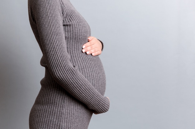 Close up of pregnant woman in gray dress holding her belly at gray background. Motherhood concept. Copy space