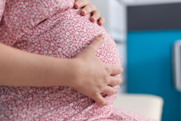 Close up of pregnant woman having hands on baby bump in medical office. Pregnant patient expecting child and thinking about parenthood. Adult with baby preparing to give birth