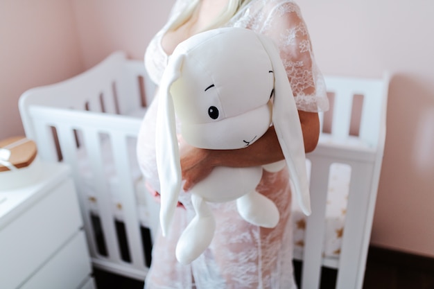 Photo close up of pregnant woman holding bunny toy while standing in baby room.
