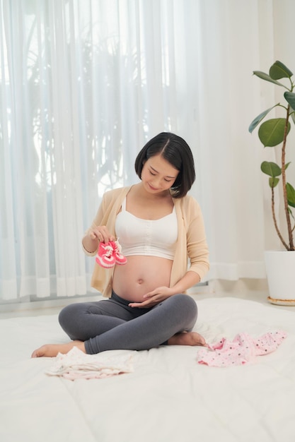 Close up of pregnant woman in home clothing holding shoes for a baby girl against her abdomen