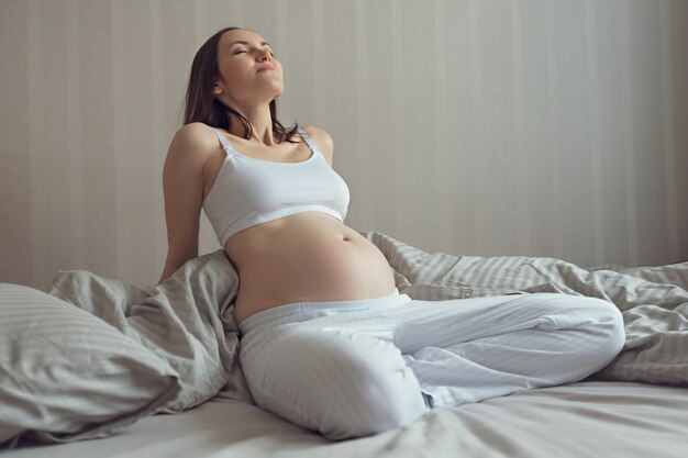 Close up of pregnant woman sitting on bed relaxing in white top and blue pants