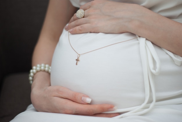 Close up of pregnant woman in white dress holding golden cross on her belly on the dark background.