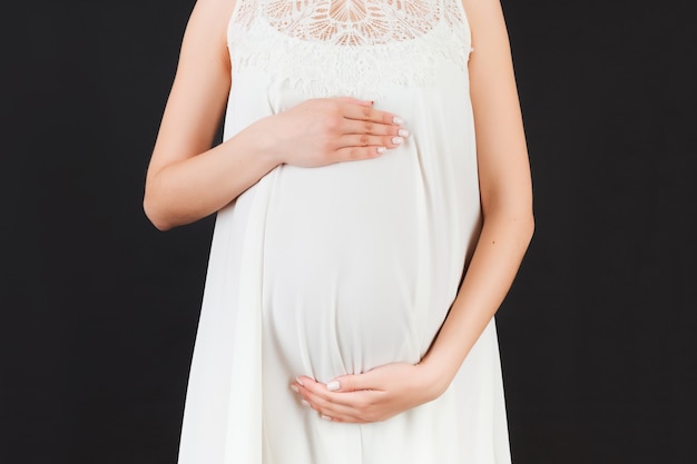 Close up of pregnant woman in white dress holding her belly at black background. Motherhood concept. Copy space.