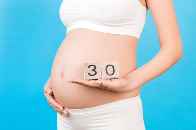 Close up of pregnant woman in white underwear holding cubes with numbers of pregnancy weeks. Thirty weeks of pregnancy period. Copy space.