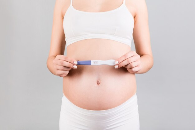 Close up of pregnant woman in white underwear holding positive pregnancy test at gray background. Motherhood concept. Copy space.