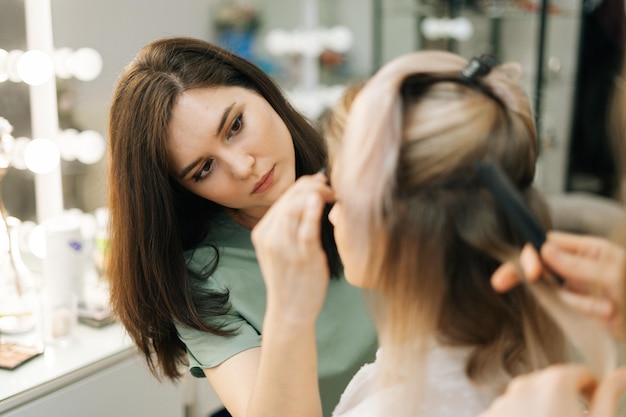 Primo piano del processo di preparazione della splendida giovane donna, il parrucchiere sta torcendo lunghi capelli biondi, truccatrice che lavora nello spogliatoio. concetto di lavoro dietro le quinte.