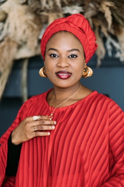 Close up of pretty african woman in red outfit and shawl on head on dark studio background. Beautiful female model with red cloth wrapped on head looking at camera.