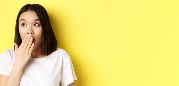 Close up of pretty asian girl looking shocked gasping and covering mouth with hand stare left at person