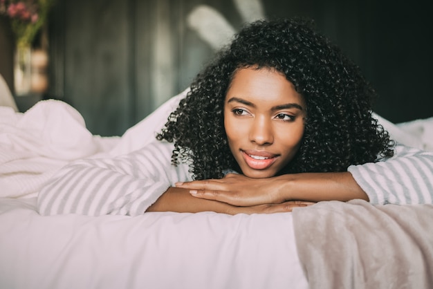 Close up of a pretty black woman with curly hair smiling and lying on bed looking away