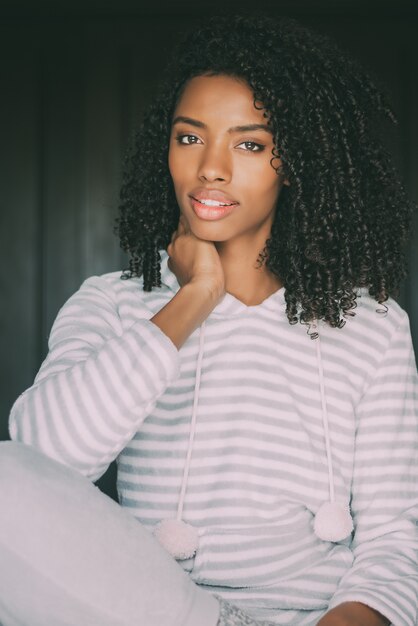 Close up of a pretty black woman with curly hair smiling sit on bed looking at the camera