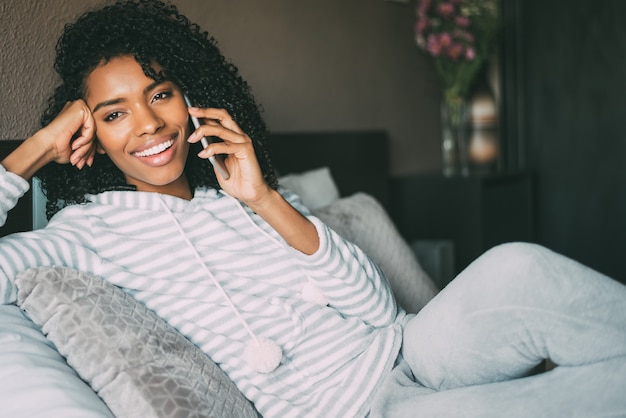 Close up of a pretty black woman with curly hair smiling and using phone on bed looking away