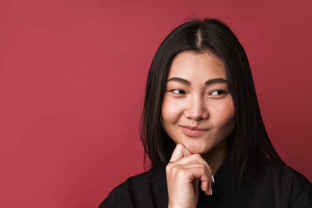 Foto primo piano di una giovane donna asiatica piuttosto pensierosa che indossa abiti casual in piedi isolata sul muro rosso, guardando lontano