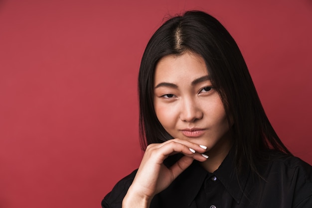 Foto primo piano di una giovane donna asiatica piuttosto pensierosa che indossa abiti casual in piedi isolata sul muro rosso, guardando la telecamera