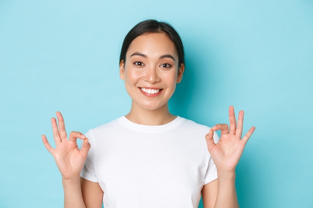 Close-up of pretty satisfied smiling asian girl in white t-shirt, showing recommendation gesture, okay sign and nod in approval, compliment choice, agree with you, standing light blue wall