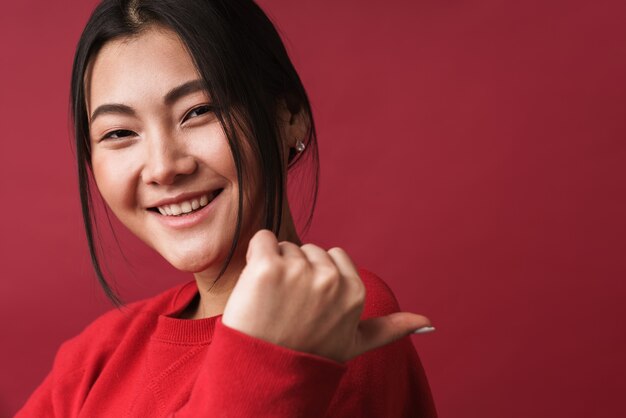 Close up of a pretty smiling young asian woman wearing casual clothes standing isolated over red wall, pointing finger away at copy space