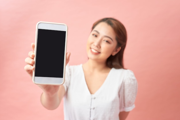 Close up of a pretty young asian woman showing blank screen mobile phone while standing and looking at camera over pink