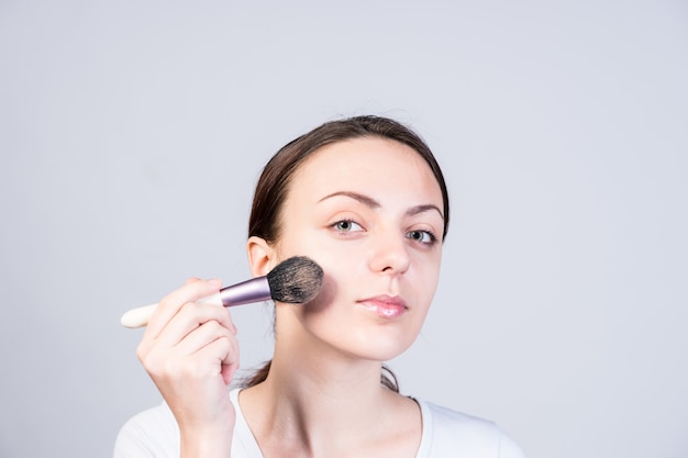 Chiuda in su abbastanza giovane donna che applica il trucco di fondazione sul viso usando un pennello mentre guarda la telecamera, isolata su uno sfondo grigio.