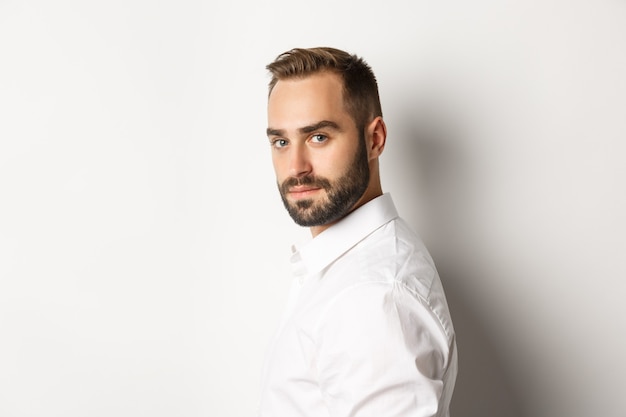 Close-up of professional business man turn face at camera, looking confident, standing against white background.