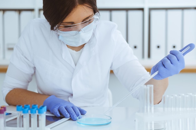 Close-up of professional female scientist in protective eyeglasses making experiment with reagents or blood test in laboratory. Medicine, biotechnology and research concept.