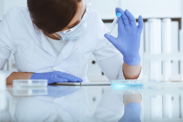 Close-up of professional female scientist in protective eyeglasses making experiment with reagents or blood test in laboratory. Medicine, biotechnology and research concept.