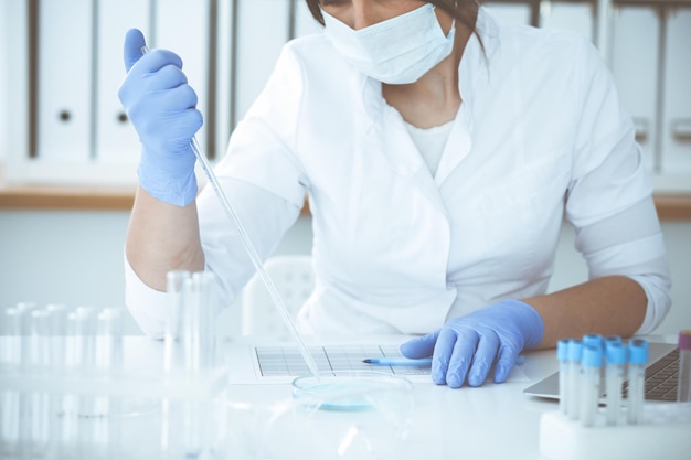 Close-up of professional female scientist in protective eyeglasses making experiment with reagents or blood test in laboratory. Medicine, biotechnology and research concept.