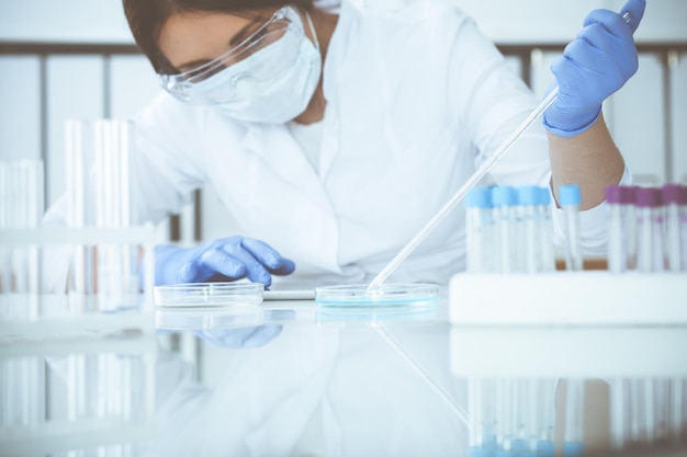 Close-up of professional female scientist in protective eyeglasses making experiment with reagents or blood test in laboratory. Medicine, biotechnology and research concept.